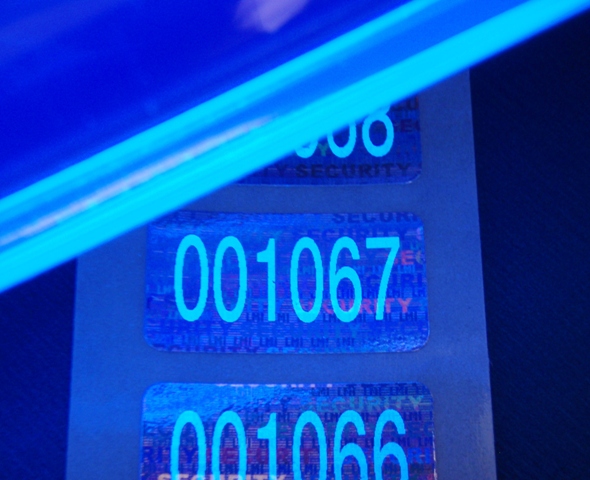 Spezielles UV-Licht macht die Sicherheitsnummerierung sichtbar Hologrammsiegel mit Lumineszenzdruck, der erst unter speziellem UV-Licht sichtbar ist